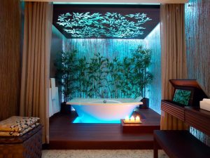 A brown bathroom with a stand alone white bath. Behind the bath is a line of greenery