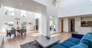 A spacious, sunlit living room with high ceilings, featuring a blue sofa and modern furniture. Large windows and white walls create an open, airy ambiance.