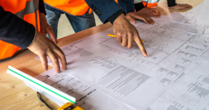 A group of people in orange vests and suits closely examine blueprints on a table. One points at details, conveying focus and collaboration.